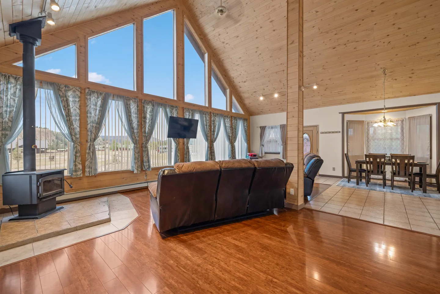 Living room with wood-paneled vaulted ceiling, large wall of A-frame windows, wood-burning stove, and brown reclining sofa.