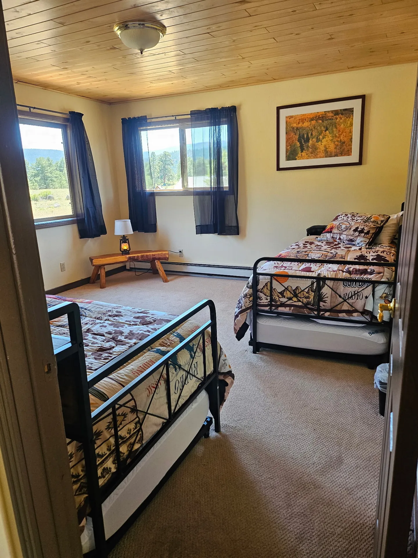Bedroom with two twin metal-frame beds, wood-paneled ceiling, and windows overlooking scenic outdoor landscape.