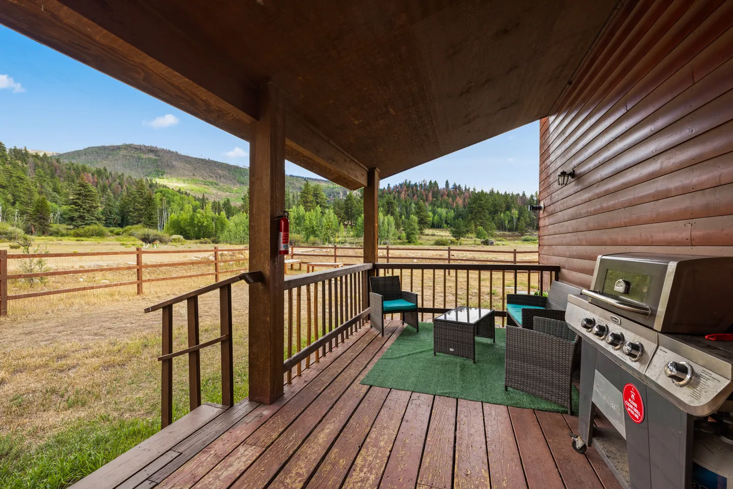 Covered wooden deck with outdoor seating, a barbecue grill, and views of a fenced field and forested hills.