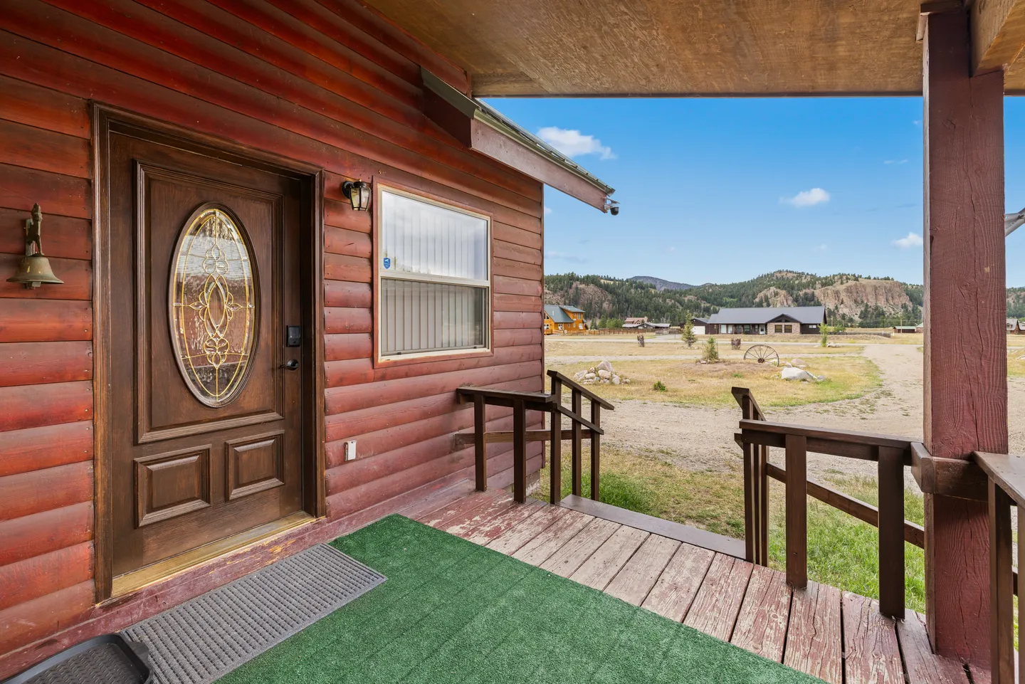 Front porch of a log cabin featuring a wooden door with an oval decorative glass insert, covered deck, and view of the open landscape.