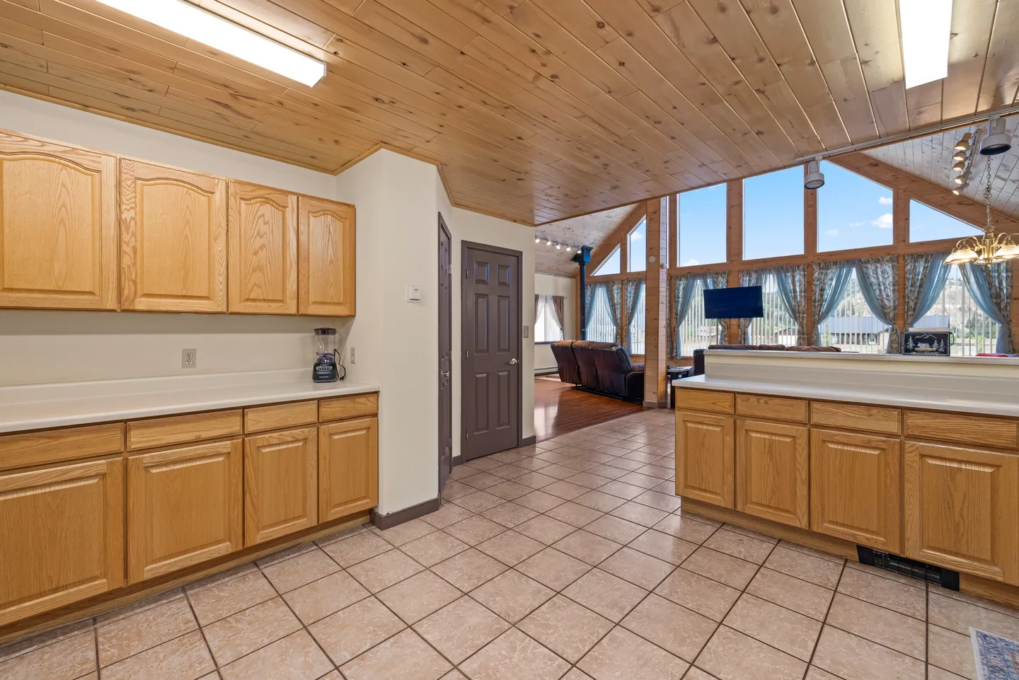 Kitchen with wooden cabinets and tile flooring leading to an open living room with vaulted wood-paneled ceilings and large windows.