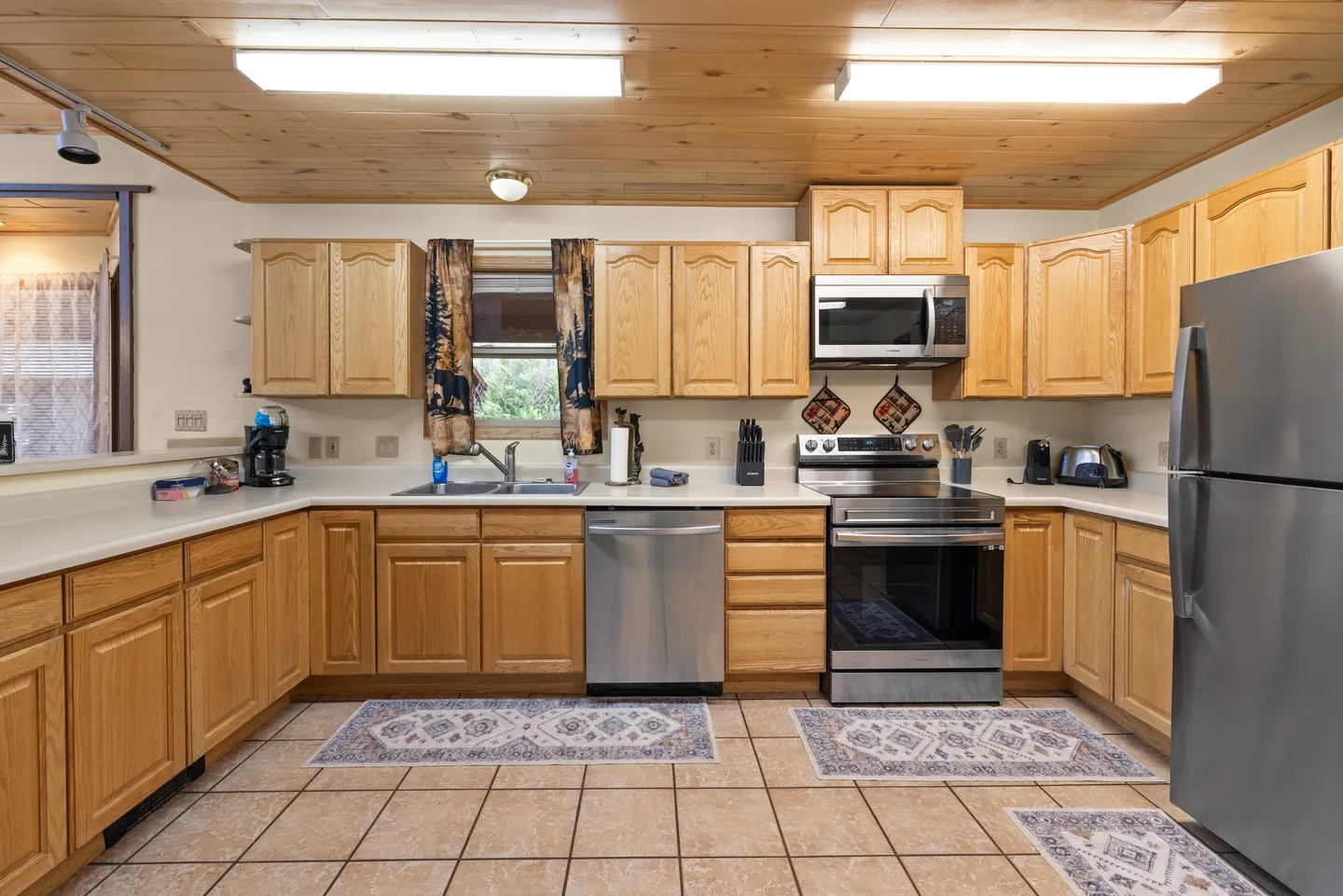 Kitchen with light wood cabinets, wood-paneled ceiling, stainless steel appliances, and tiled flooring.