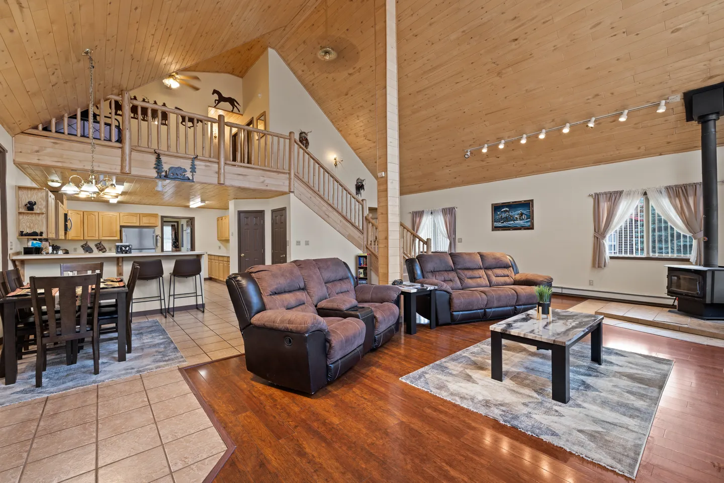 Open-concept living room and kitchen featuring vaulted wood-paneled ceilings, a loft overlooking the main floor, brown reclining sofas, and a wood stove.