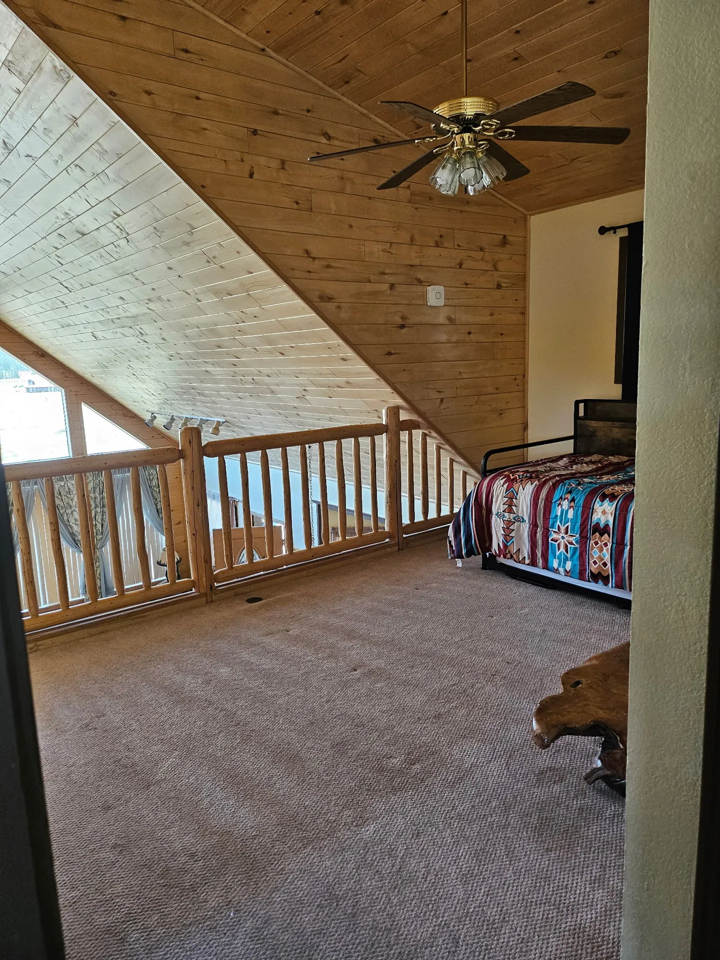 Loft bedroom area with carpeted floors, wood railings overlooking lower level, wood-paneled angled ceiling, and a bed with a patterned quilt.