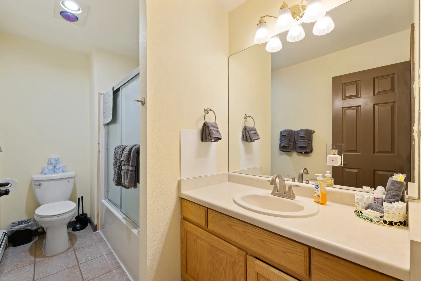 Bathroom with light wood vanity, large mirror, toilet, and bathtub with sliding glass doors.