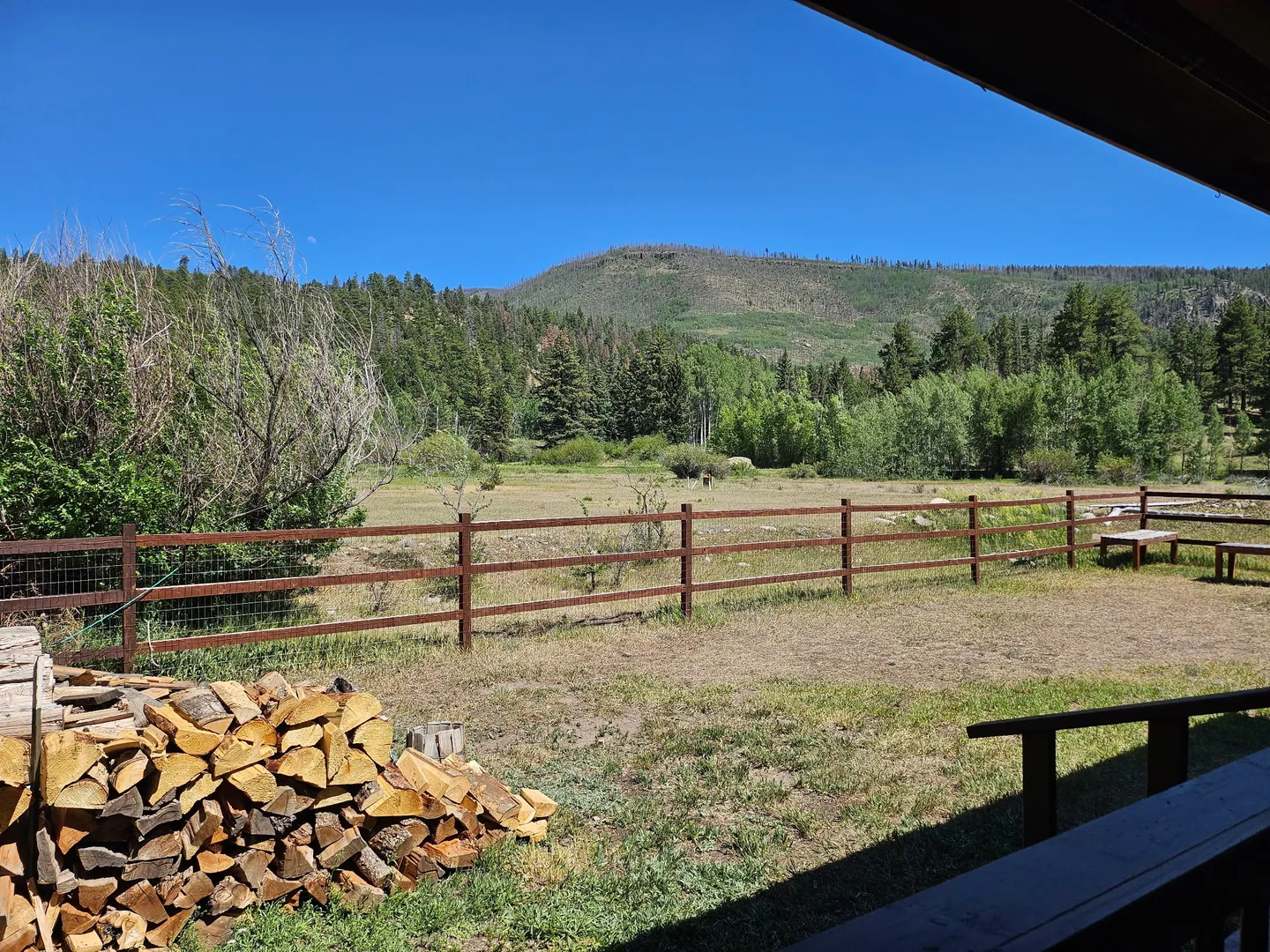 View from a covered porch looking out over a fenced yard with a woodpile toward open fields and tree-covered hills under a clear blue sky.