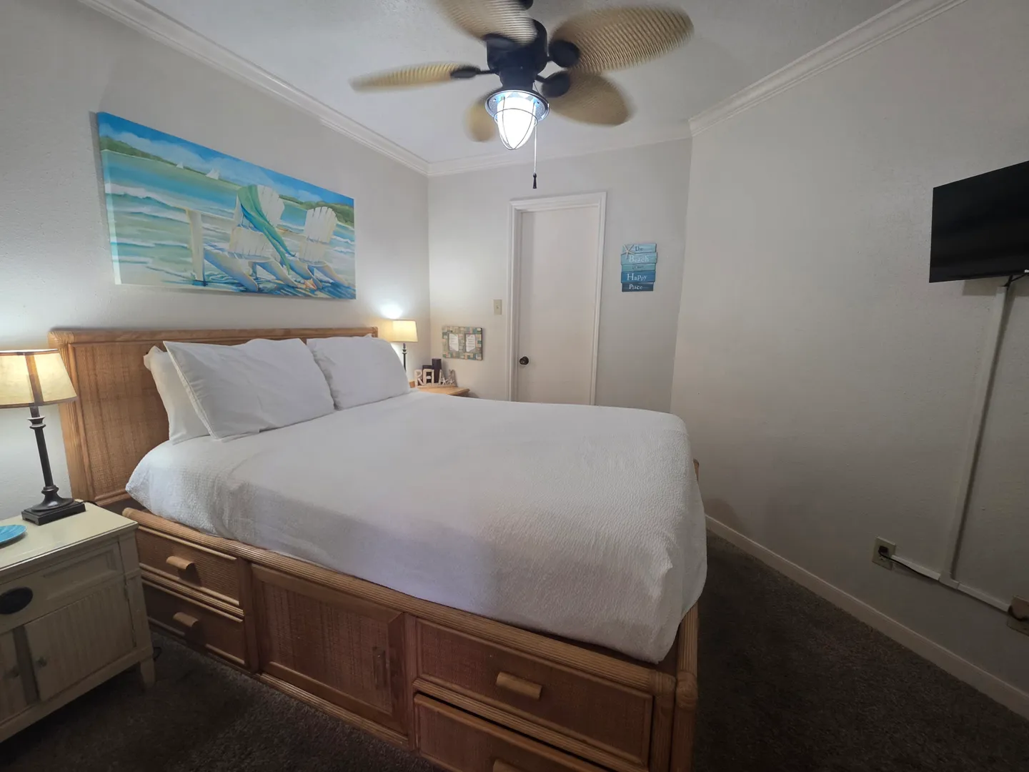 Bedroom with a white-dressed bed featuring built-in storage drawers, bedside tables with lamps, a ceiling fan, and a wall-mounted TV.