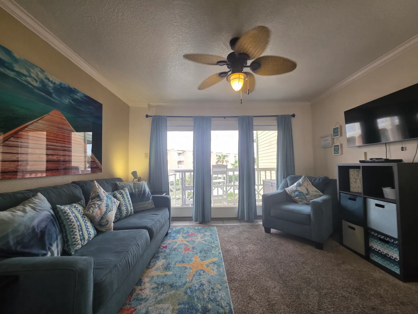 Living room with a blue sofa, matching armchair, ceiling fan, and sliding glass doors leading to a balcony.