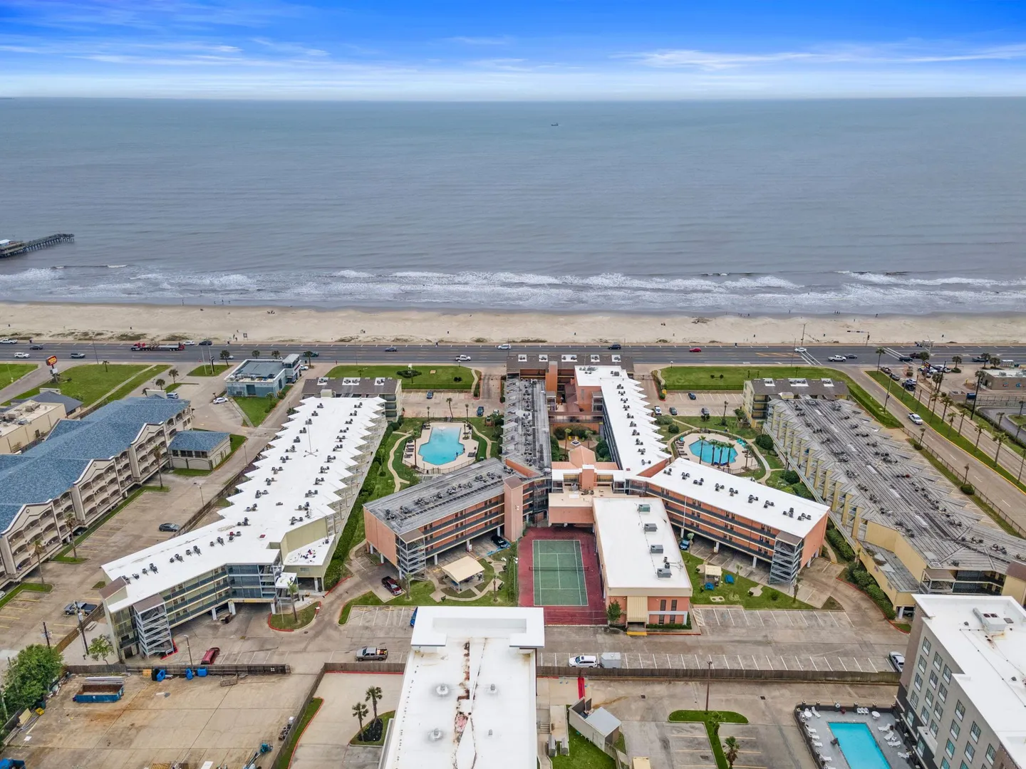 Aerial view of a beachfront condominium complex featuring swimming pools, a tennis court, parking lots, and the ocean coastline.