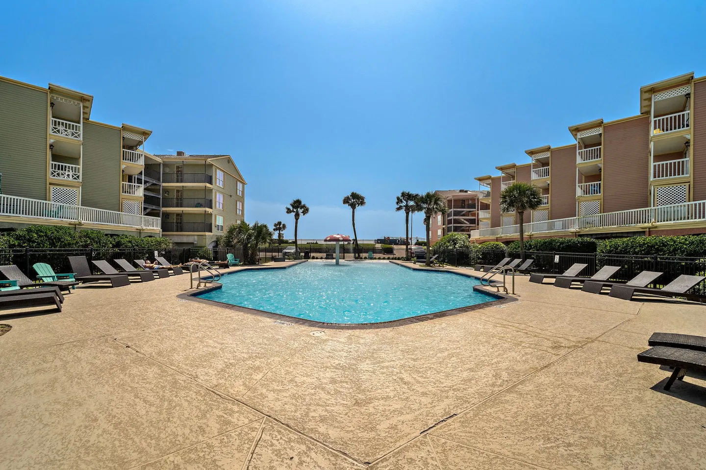 Outdoor swimming pool surrounded by lounge chairs and multi-story apartment buildings under a clear blue sky.
