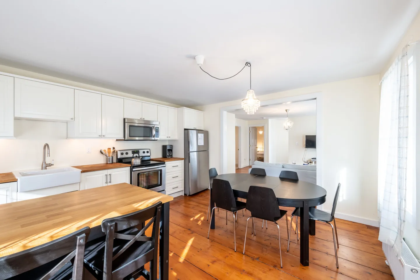 Open-concept kitchen and dining area with wood plank flooring, white shaker cabinets, an island with seating, a black dining table, and a wide doorway to the living room.