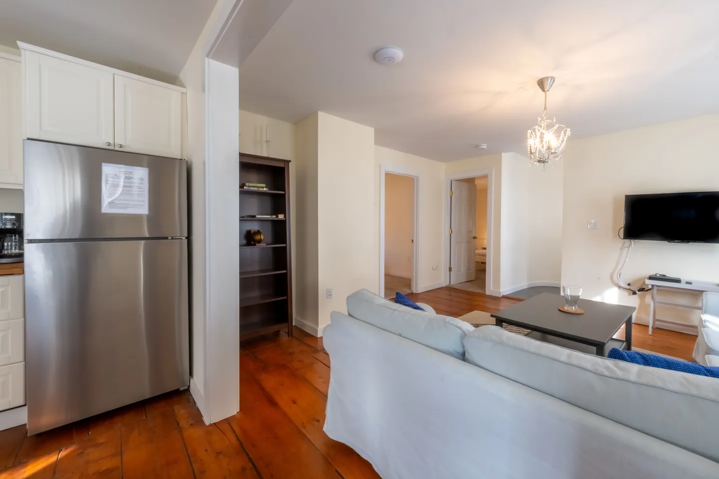 Open living area with hardwood floors, a light-colored sofa, coffee table, wall-mounted television, and a partial view of the kitchen with a stainless steel refrigerator.