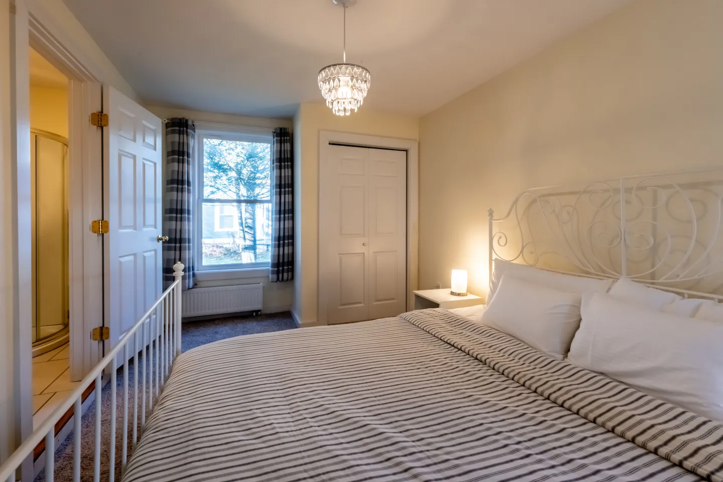 Bedroom with a white metal bed, chandelier, window with plaid curtains, and an open door showing an adjoining bathroom.