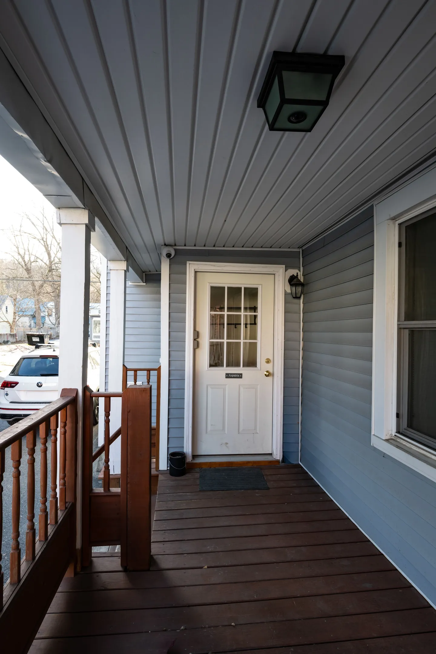 A covered porch with wood plank flooring, grey siding, and a white entrance door with glass panes.