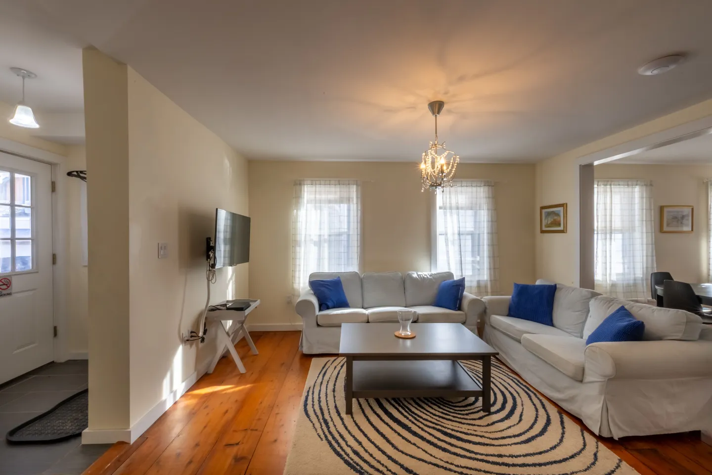 Living room with white slipcovered couches, a coffee table on a patterned rug, wood floors, and a wall-mounted TV.