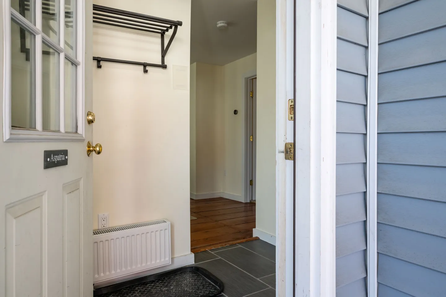 Open white entry door looking into an entryway with dark tiled floor, a wall rack, and hardwood flooring in the hallway beyond.