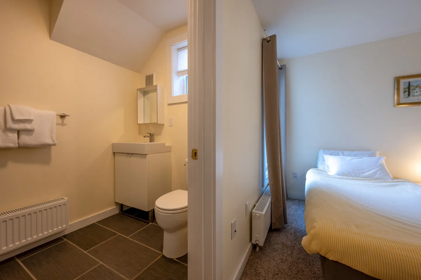 A view showing a bedroom on the right with a single bed and an adjoining bathroom on the left with a vanity, toilet, and tiled floor.