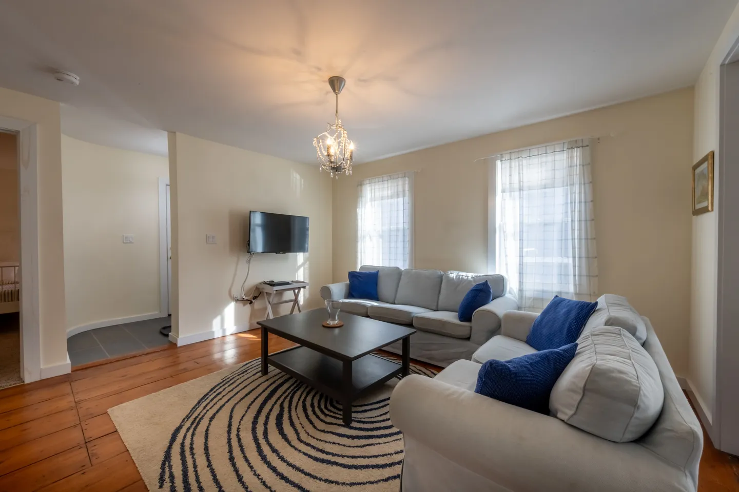 Living room with light-colored sofas, dark coffee table, patterned area rug, wall-mounted television, and a small chandelier.