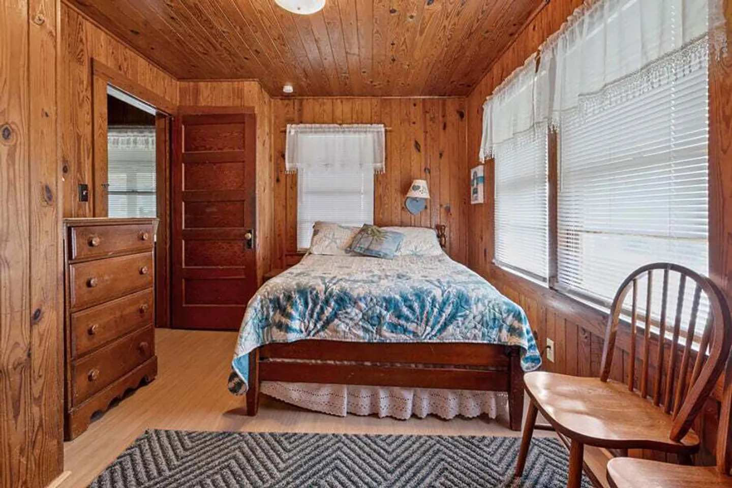 Bedroom with knotty pine wood-paneled walls and ceiling, a wooden bed, dresser, and windows with blinds.