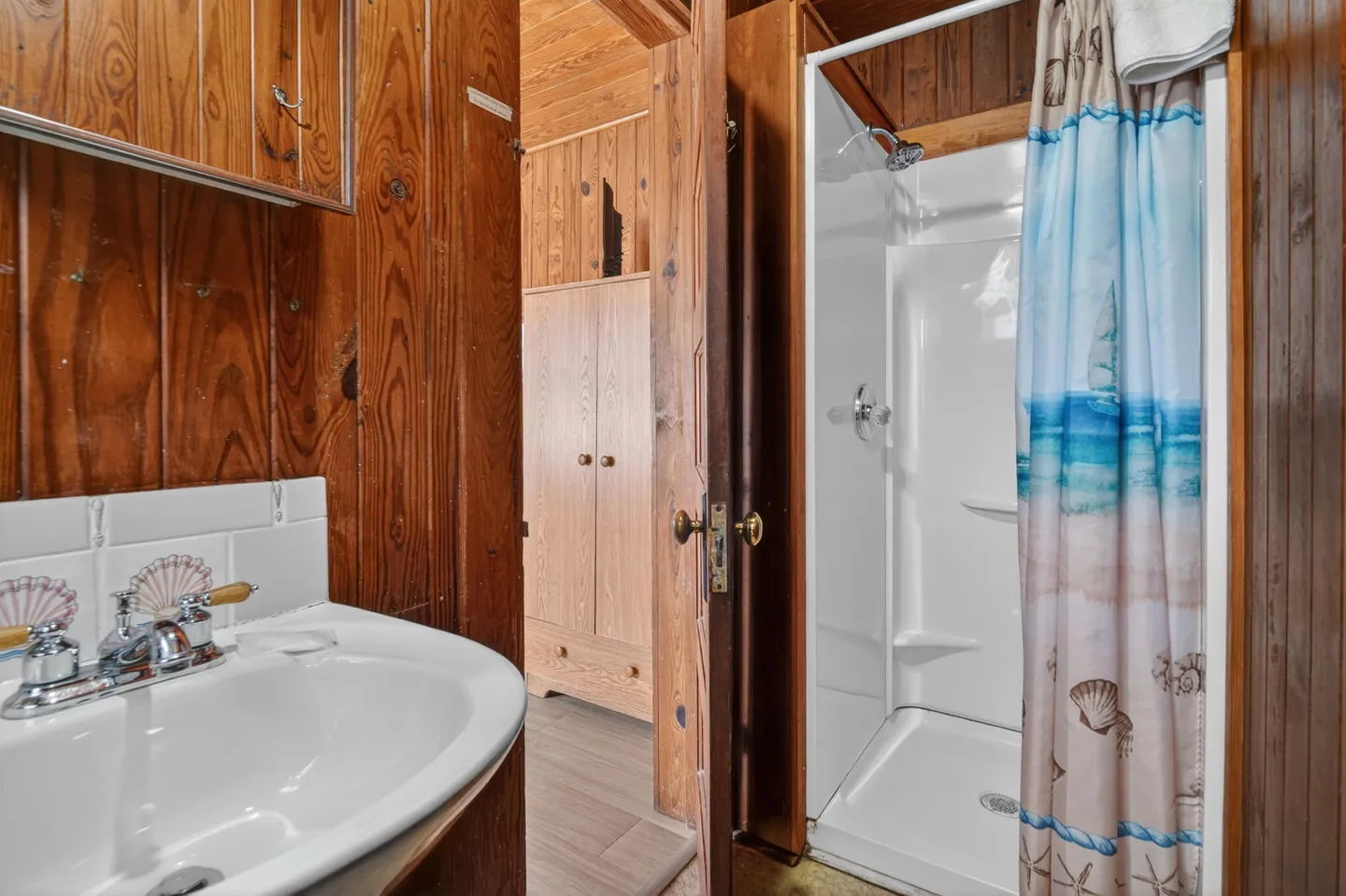 Bathroom with wood-paneled walls, white sink with seashell handles, and a walk-in shower with a beach-themed curtain next to an open doorway.