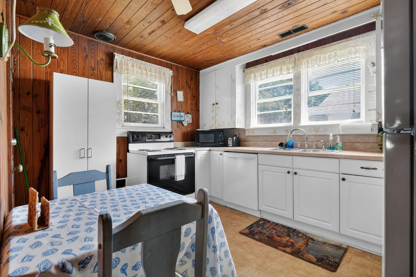 Kitchen with wood-paneled walls and ceiling, white cabinetry, dining table covered in a patterned tablecloth, and windows with blinds.