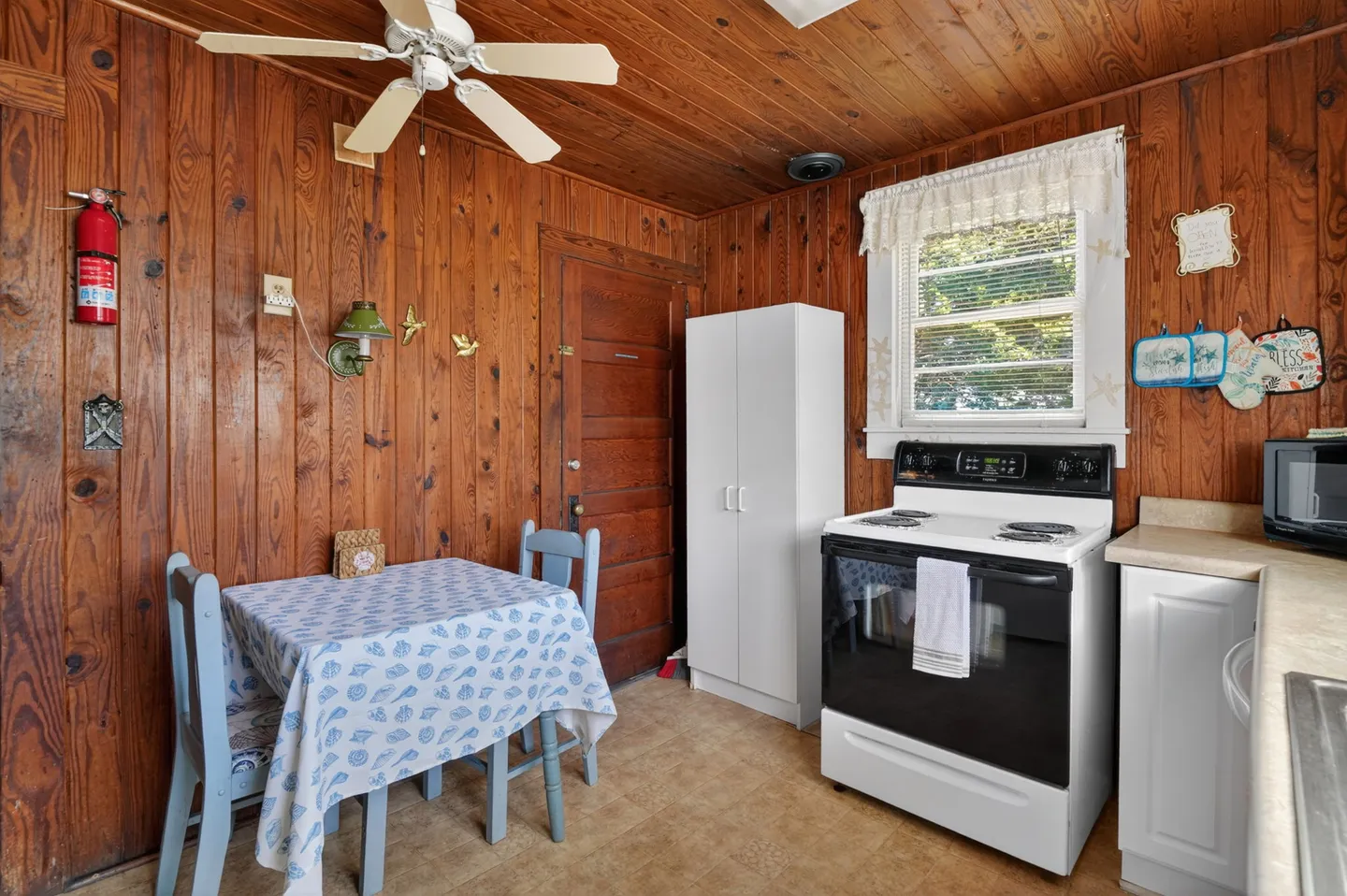 A kitchen and dining nook with wood-paneled walls, wood ceiling with a fan, a small table with chairs, an electric stove, and a window.