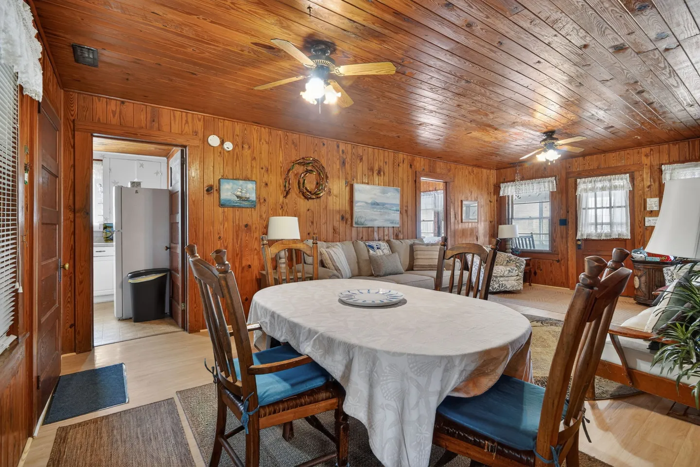 Dining area with a covered table and wooden chairs leading into a wood-paneled living room and adjacent kitchen.