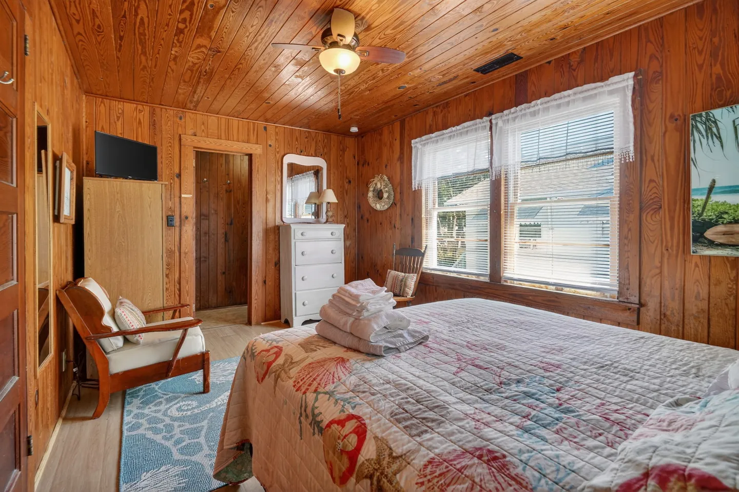 Bedroom with knotty wood-paneled walls and ceiling, a patterned bedspread, dresser, and armchair.