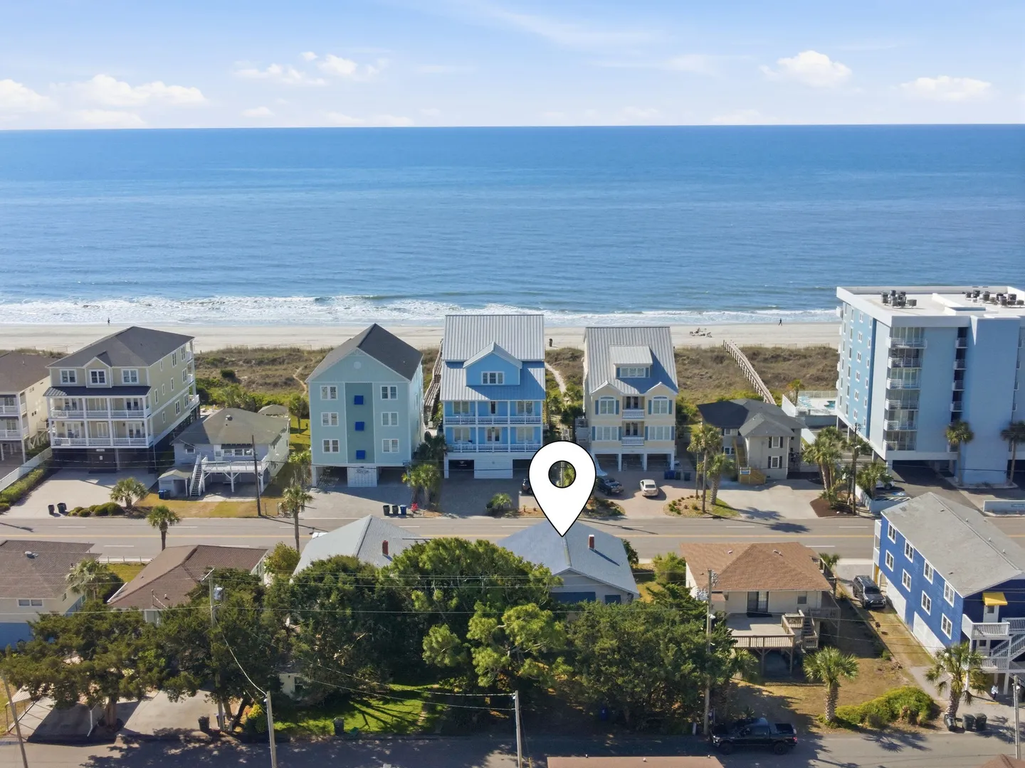 Aerial view showing coastal houses, a road, a sandy beach, and the blue ocean under a clear sky, with a location pin marker on one house.