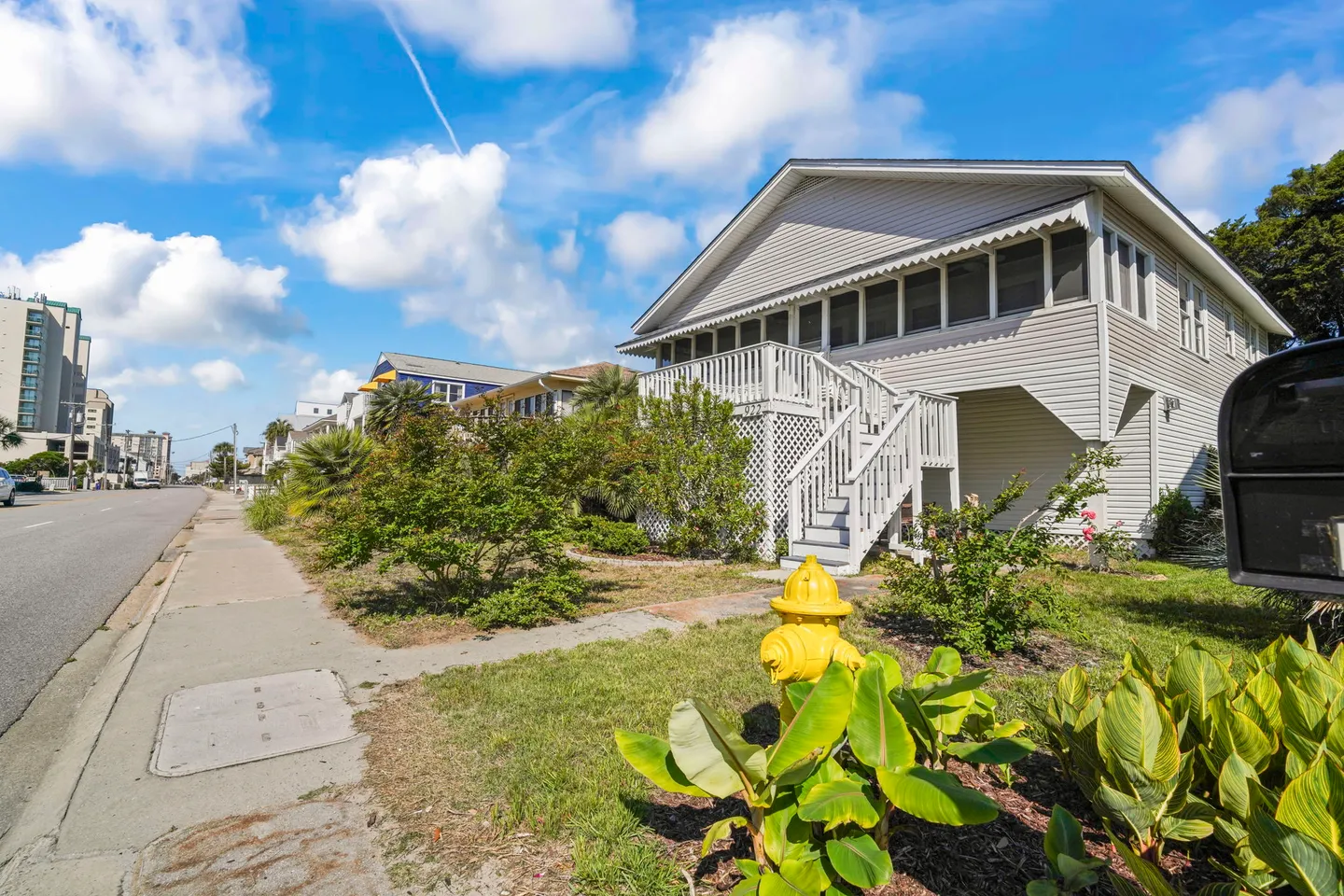 Exterior street view of an elevated coastal house with white siding, an outdoor staircase, and a sidewalk lined with greenery.