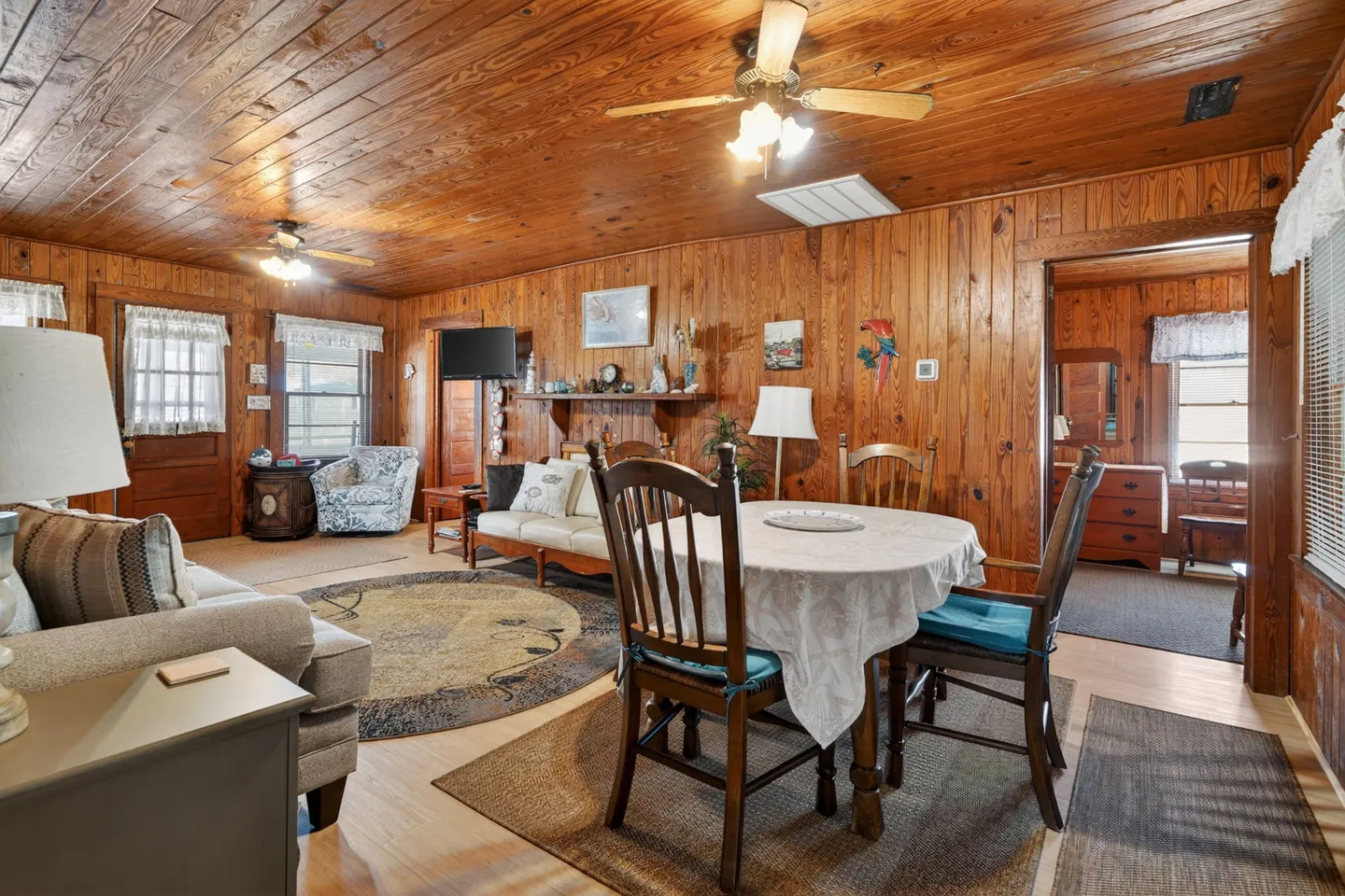 Open-concept cabin interior with knotty pine paneled walls and ceiling, featuring a small dining table and a seating area with sofas.