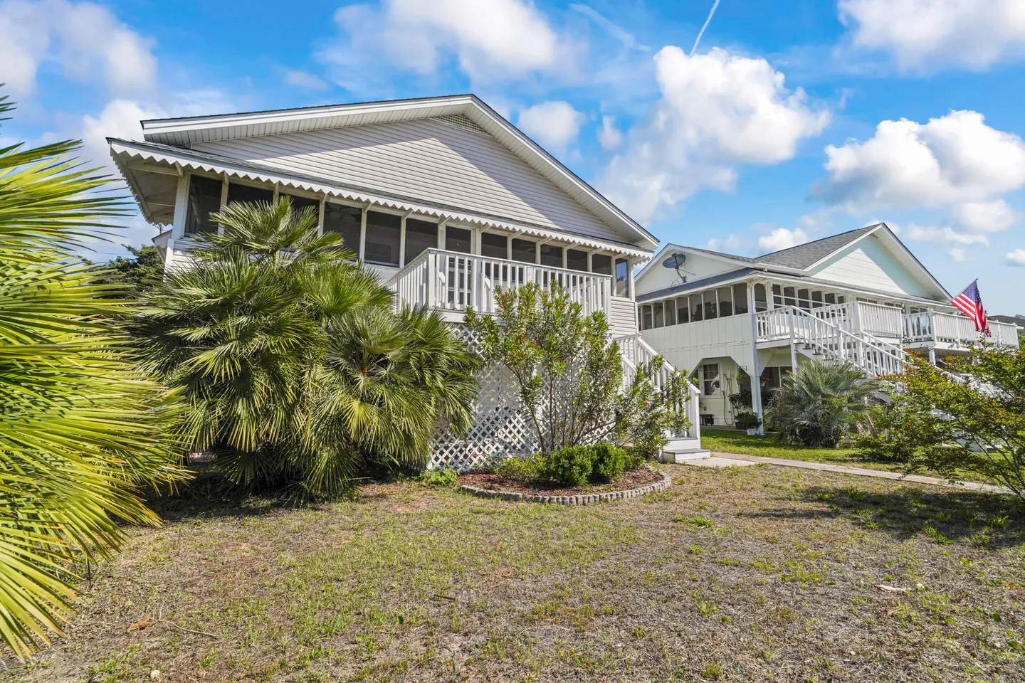 Exterior view of an elevated home with white siding, a screened porch, an open deck, and palm landscaping under a blue sky.