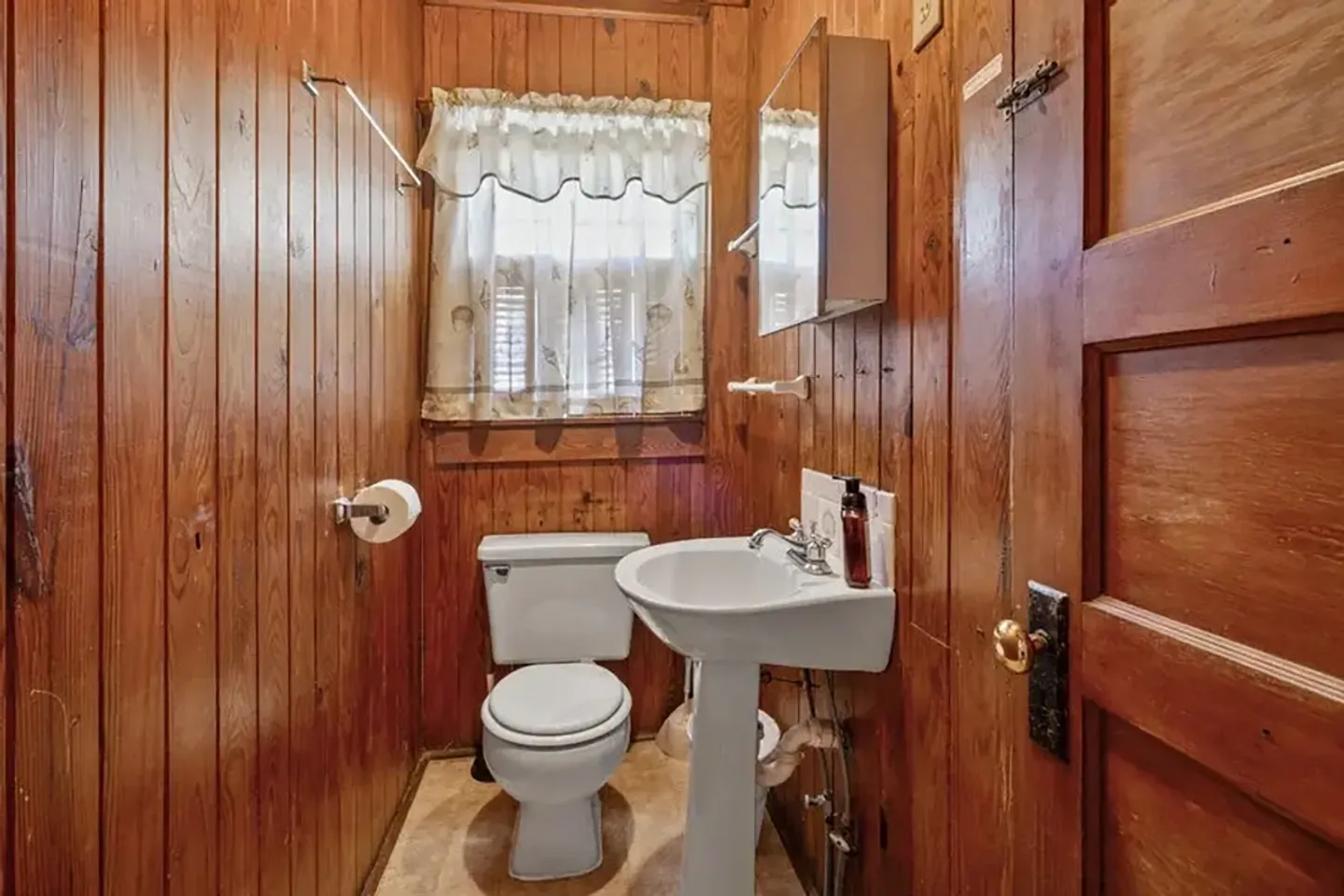 Bathroom with wood-paneled walls, a white pedestal sink, white toilet, and a small curtained window.