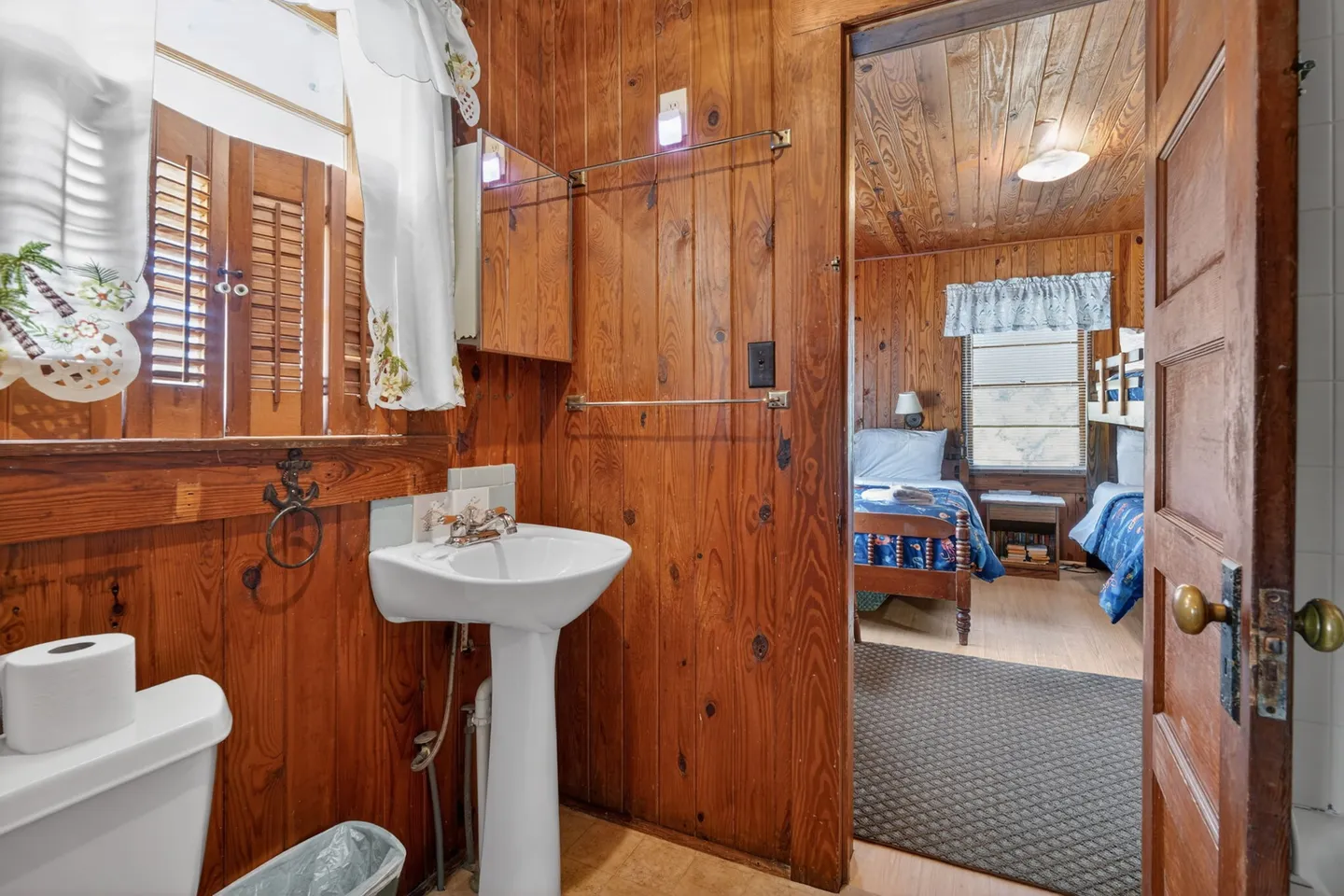 Bathroom with wood-paneled walls, pedestal sink, and toilet, looking into an adjacent wood-paneled bedroom.