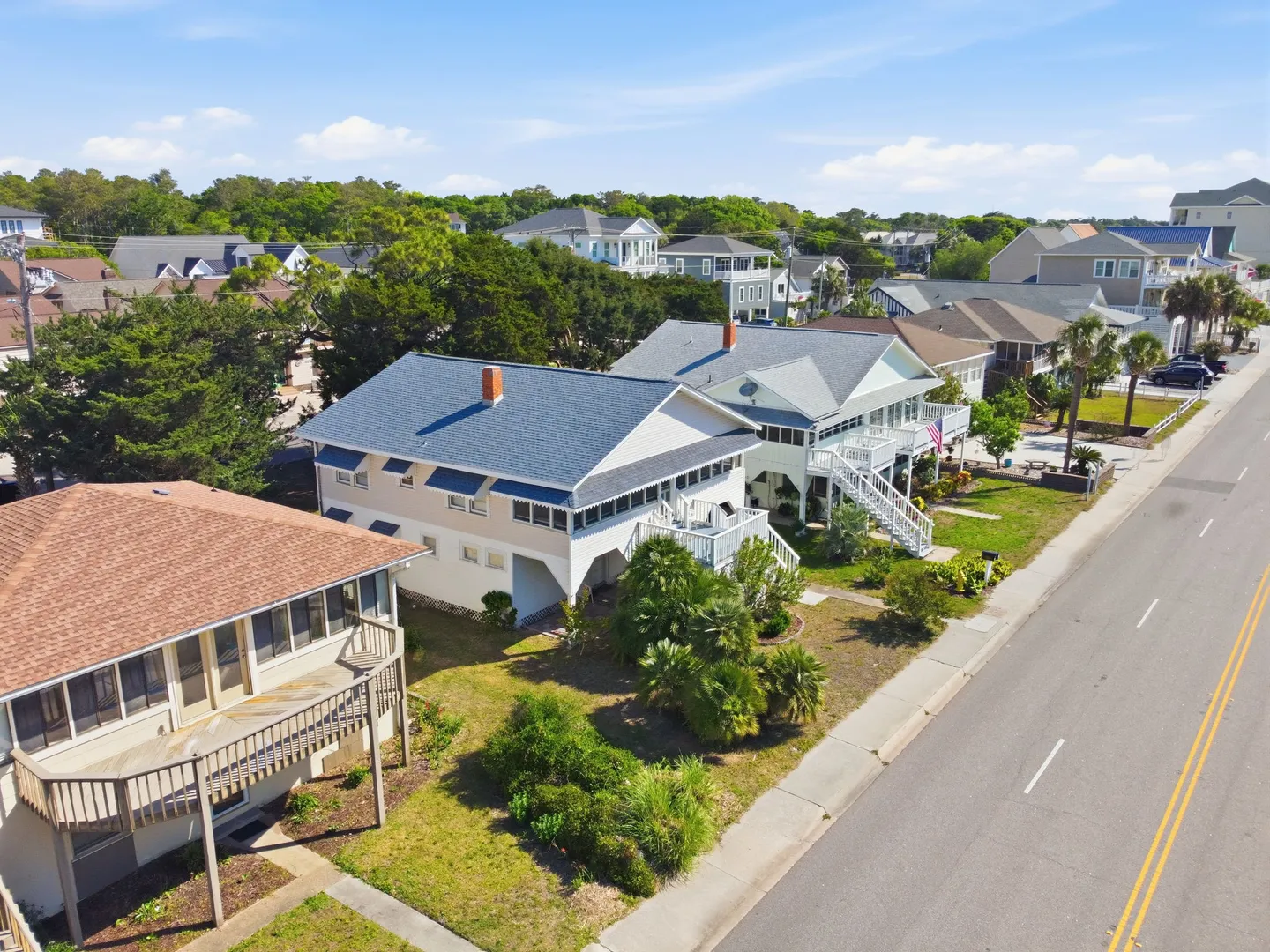 Aerial view of a coastal street lined with multi-story beach houses and lush green trees under a bright blue sky.