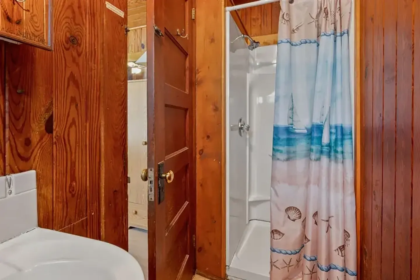 Bathroom with wood-paneled walls, an open wooden panel door, a corner of a white toilet, and a stand-up shower stall with a nautical-themed shower curtain.