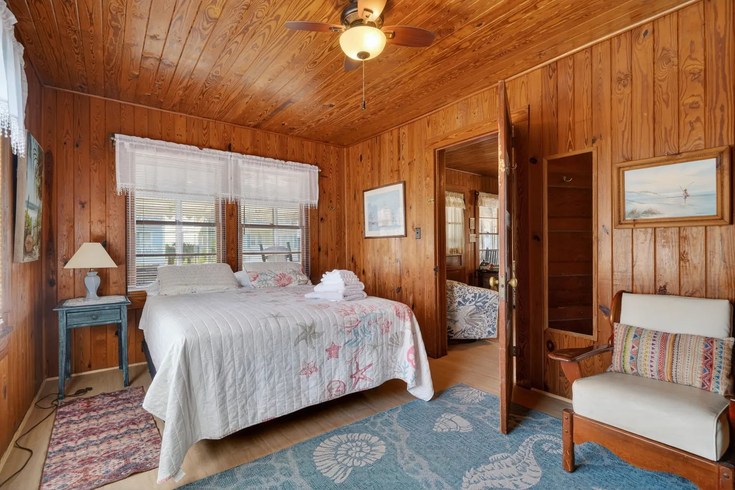 Bedroom with knotty pine wood-paneled walls and ceiling, featuring a bed made with a starfish-patterned quilt, an armchair, and a doorway showing an adjoining room.