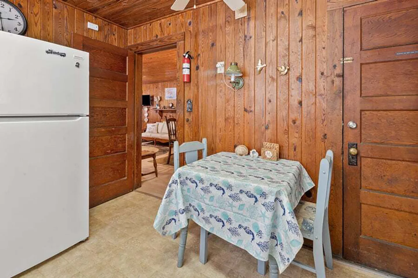 Dining nook with a small table covered in a patterned tablecloth and wood-paneled walls next to a refrigerator.