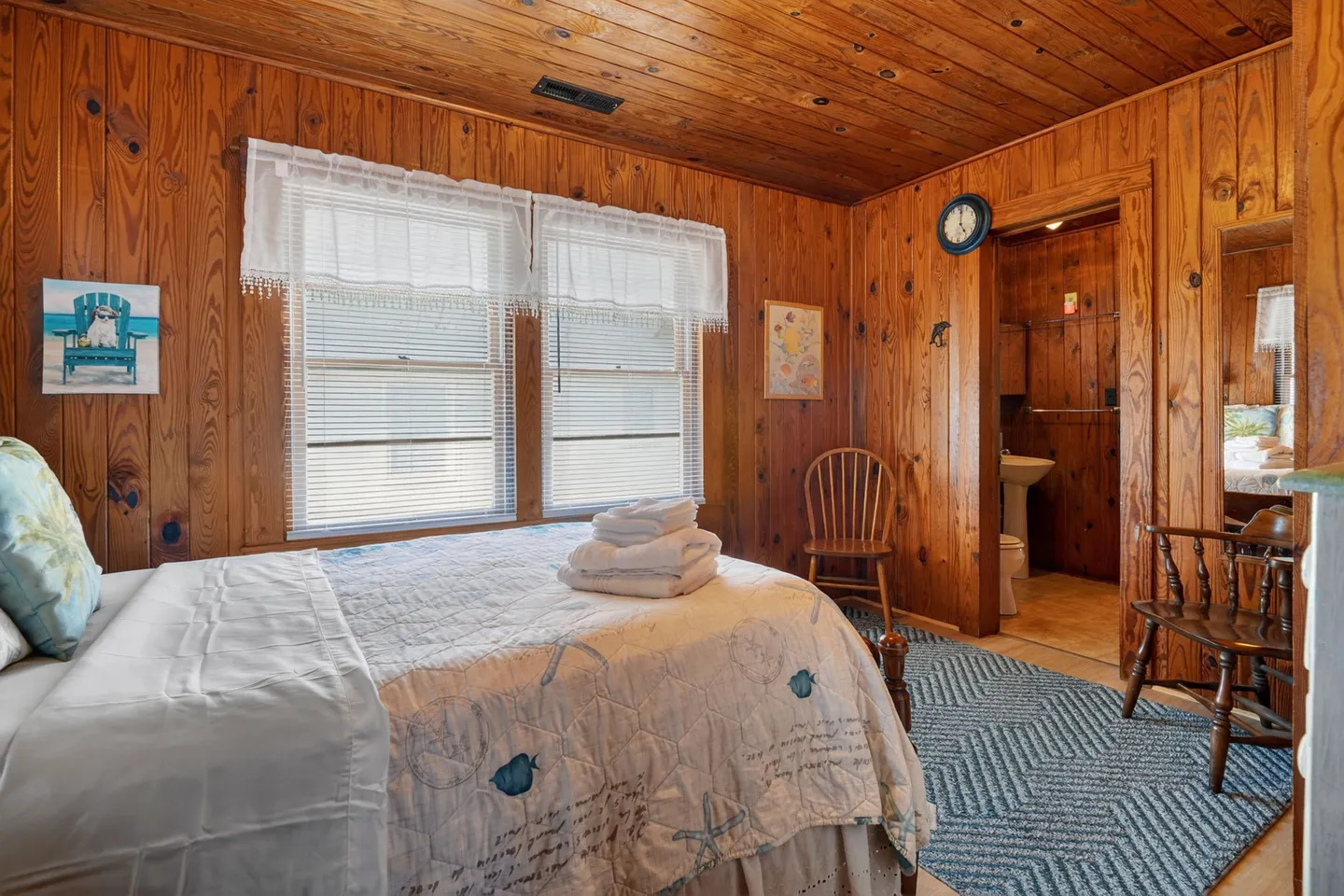 Bedroom with knotty pine wood-paneled walls and ceiling, featuring a bed made with a nautical quilt and an open doorway leading to a bathroom.