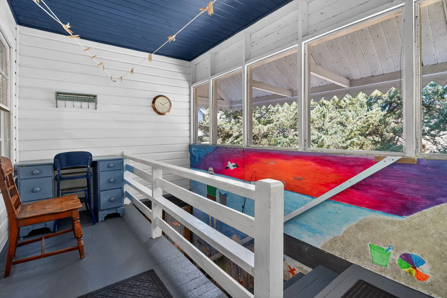 Enclosed porch or landing with white siding, blue ceiling, a small desk, and a colorful painted mural along the stair railing.