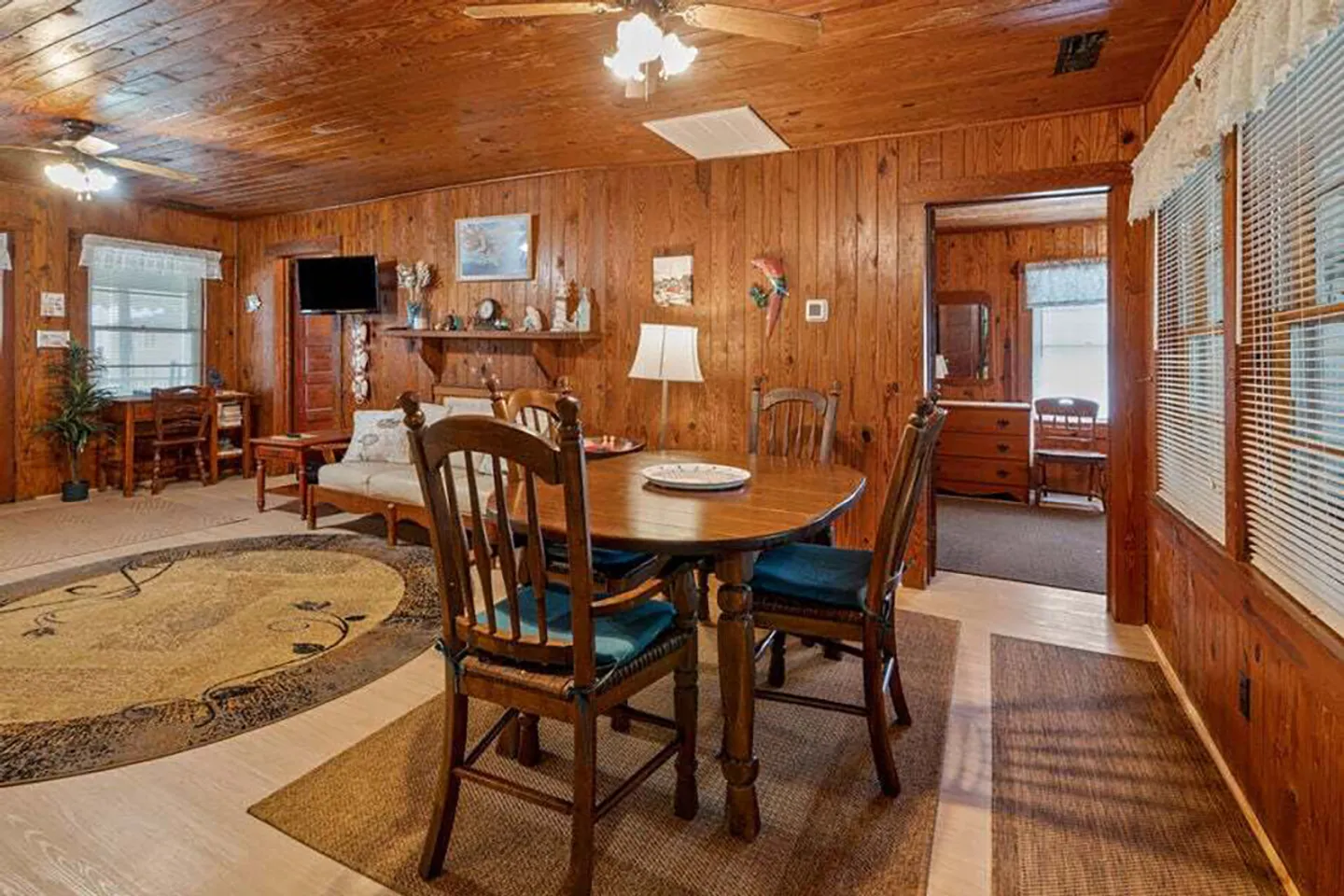 Dining area with a wooden table and chairs in a wood-paneled room opening to a living area and doorway to a bedroom.