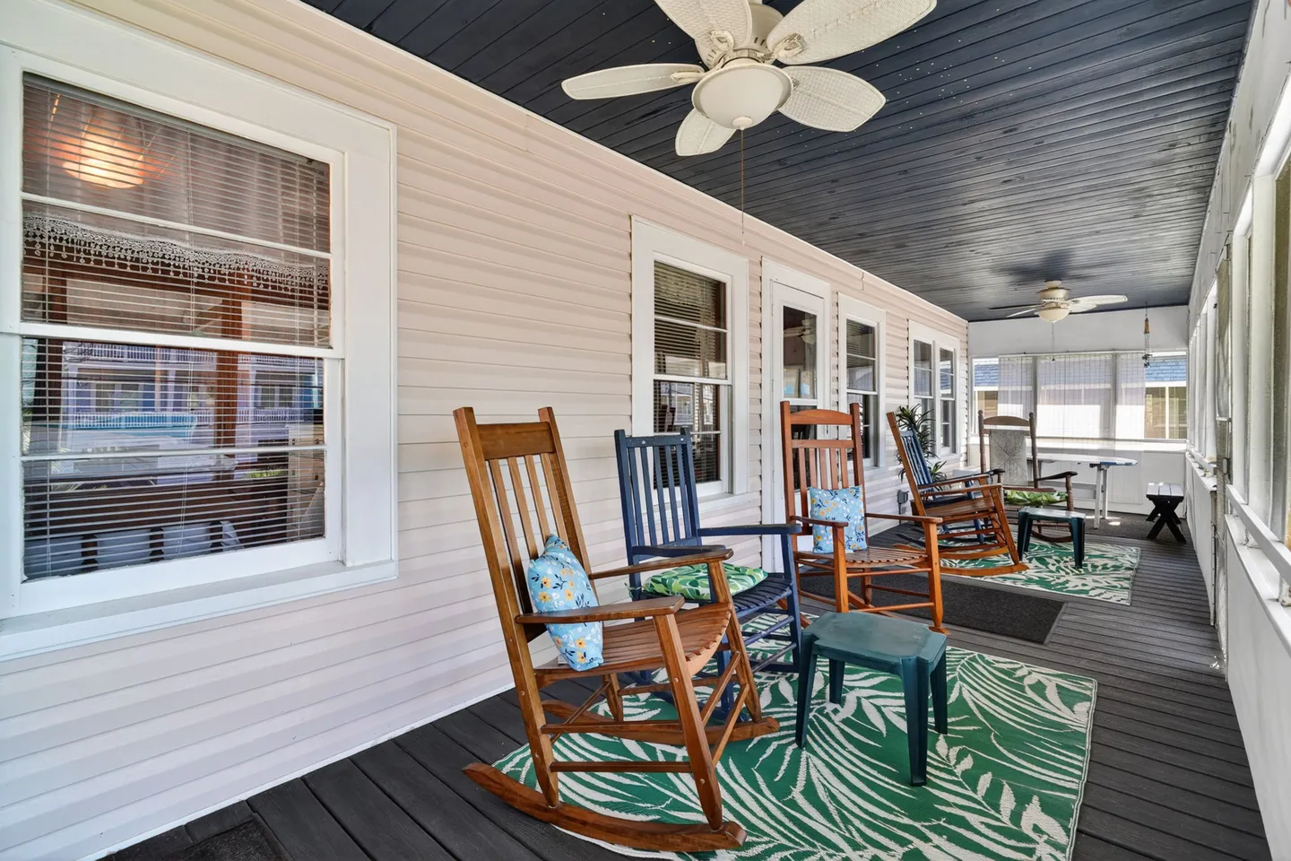 Screened porch furnished with rocking chairs on green patterned rugs beneath ceiling fans and a dark wood-paneled ceiling.