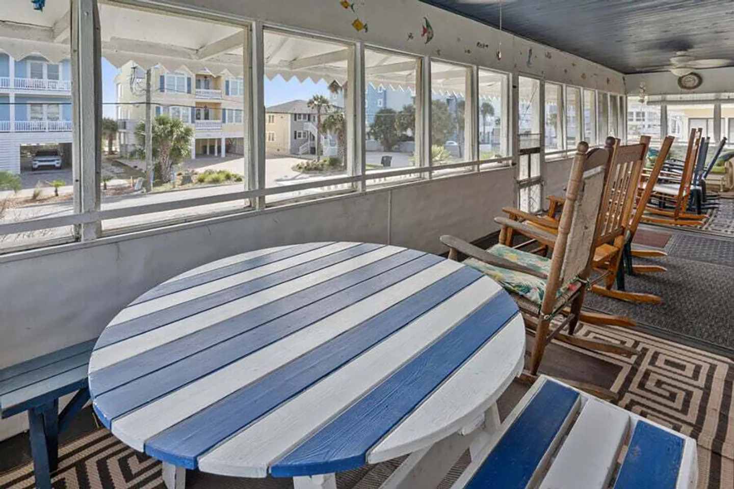 Screened porch furnished with a blue-and-white striped round picnic table and a row of wooden rocking chairs overlooking the neighborhood.