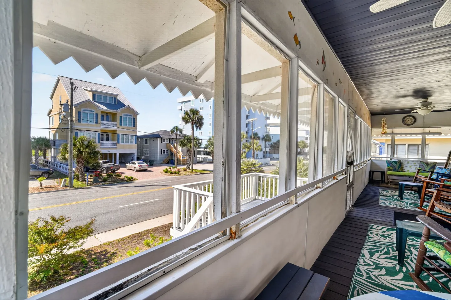 Screened porch overlooking a coastal neighborhood street with rocking chairs and area rugs on dark wood flooring.