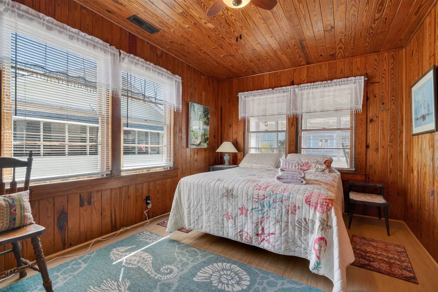 Bedroom with knotty wood-paneled walls and ceiling, a bed with coastal seashell-patterned quilt, and windows with blinds and valances.