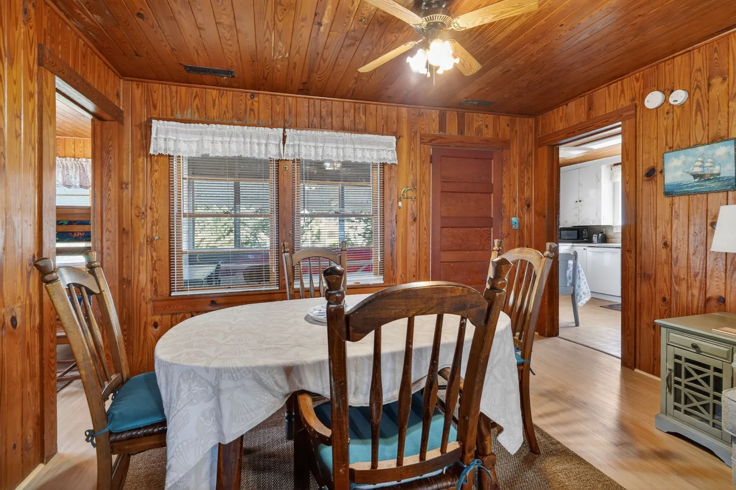 Dining area with wood paneled walls, ceiling fan, and dining table with chairs under a white tablecloth.