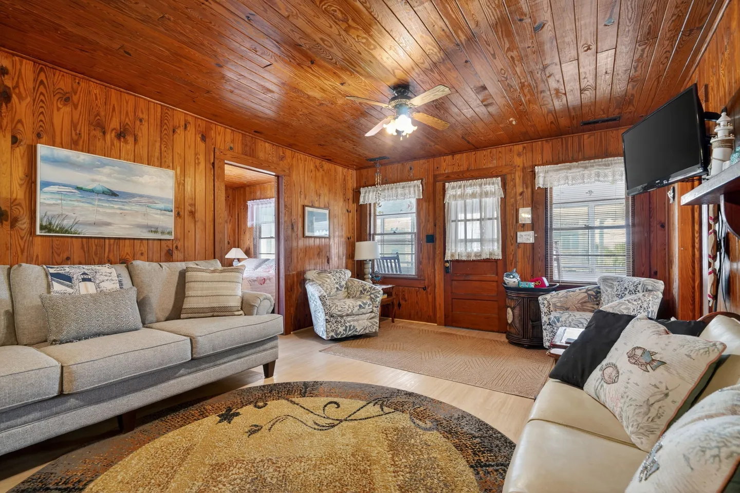 Living room with knotty wood-paneled walls and ceiling, furnished with a sofa, patterned armchairs, and area rugs.