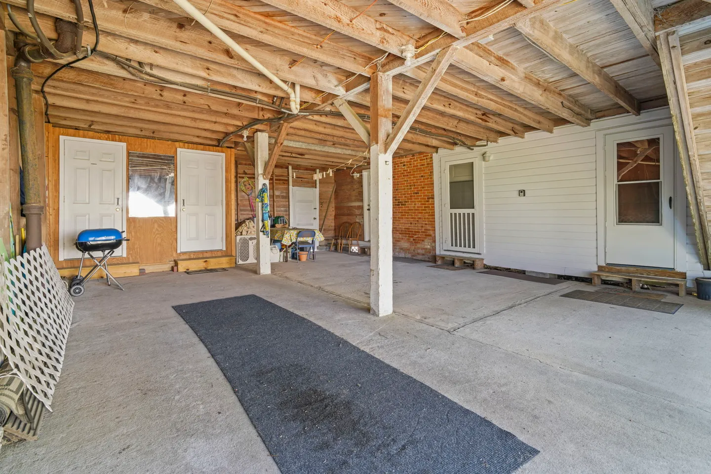 Open covered carport or under-deck patio with concrete flooring, exposed wooden ceiling beams, support posts, and multiple doors leading to the building.