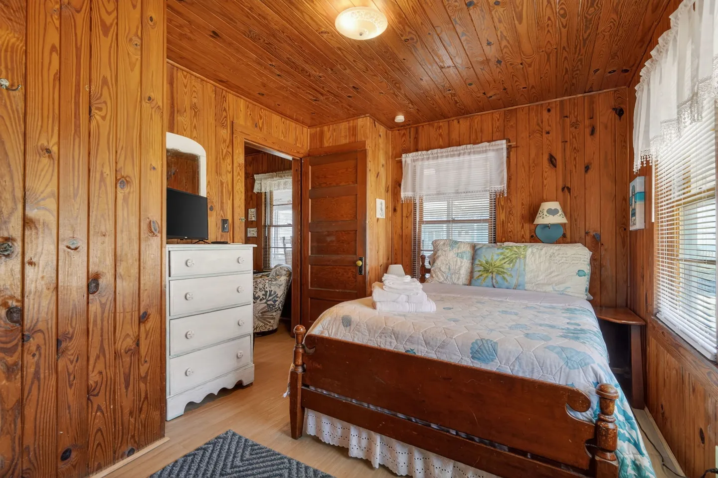 Bedroom with wood-paneled walls and ceiling, a wooden bed, a white dresser, and windows with white curtains.
