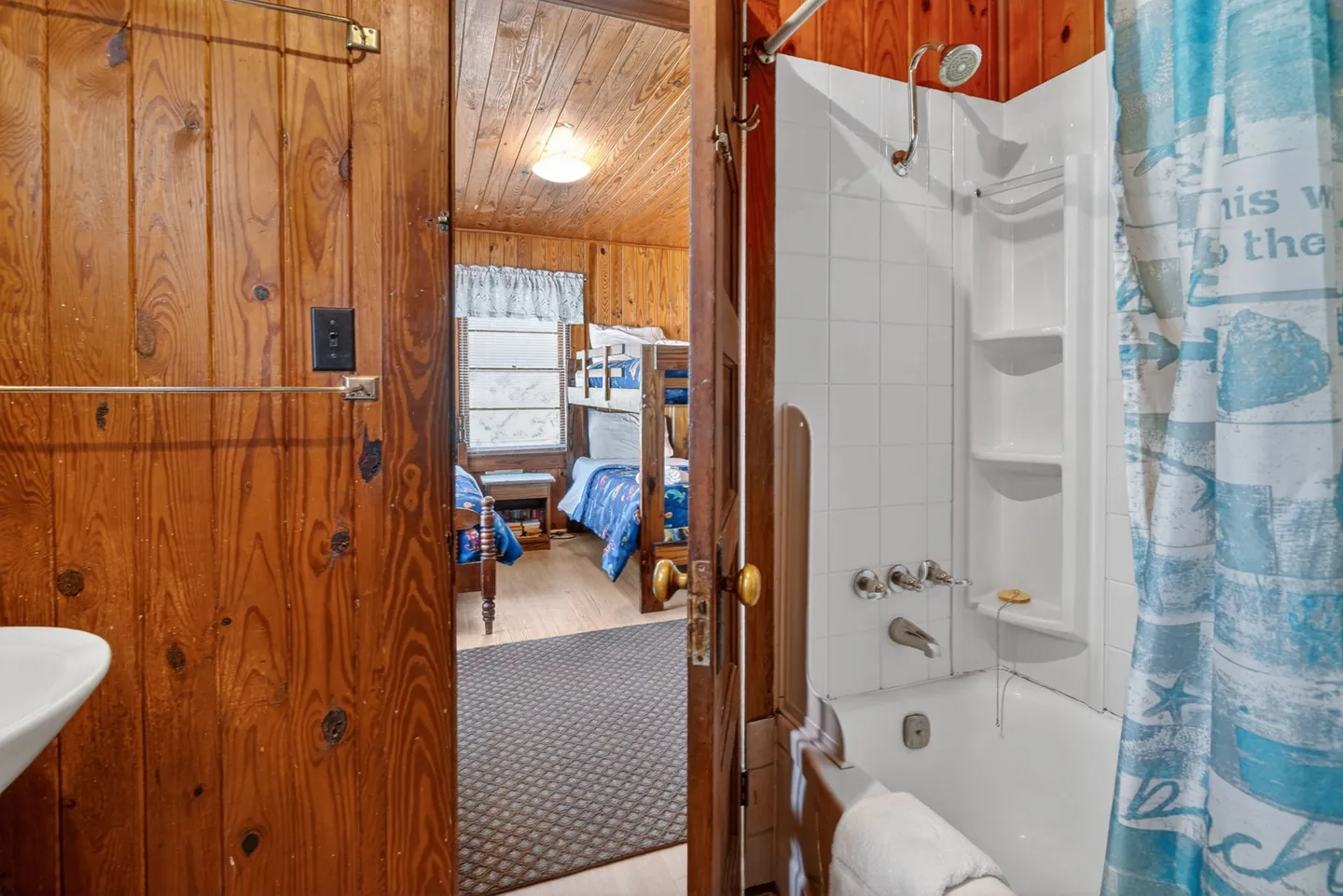 View through an open wood-paneled bathroom doorway showing a tiled bathtub and shower on the right, looking into a wood-paneled bedroom with bunk beds.